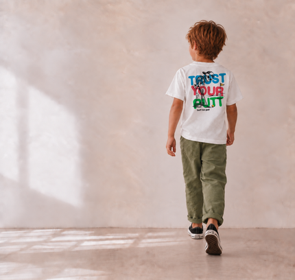 A child walking away from the camera wearing a white graphic t-shirt with 'TRUST YOUR GUT' printed in blue, red, and green on the back, paired with olive green pants and black sneakers, set against a textured beige wall with soft window shadows.