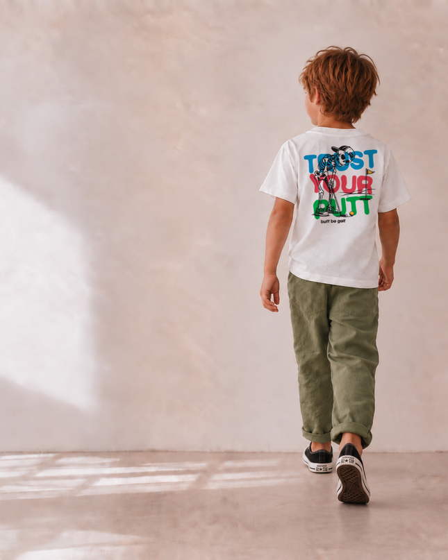 A child walking away from the camera wearing a white graphic t-shirt with 'TRUST YOUR GUT' printed in blue, red, and green on the back, paired with olive green pants and black sneakers, set against a textured beige wall with soft window shadows.