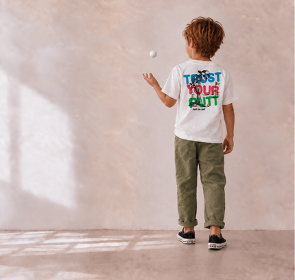 A child viewed from behind wearing a white graphic T-shirt with colorful text on the back, olive green pants, and black sneakers, standing in a minimalist room with a textured beige wall and floor while tossing a white ball.