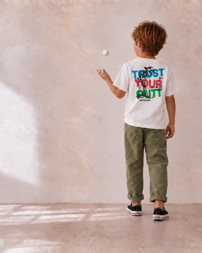 A child viewed from behind wearing a white graphic T-shirt with colorful text on the back, olive green pants, and black sneakers, standing in a minimalist room with a textured beige wall and floor while tossing a white ball.