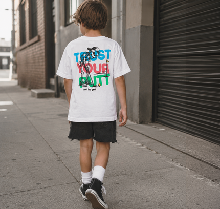 A child viewed from behind walking on a city sidewalk, wearing a white graphic T-shirt with large multicolor text and artwork on the back, black shorts, white socks, and black sneakers.