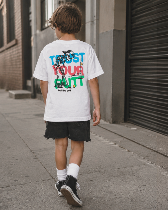 A child viewed from behind walking on a city sidewalk, wearing a white graphic T-shirt with large multicolor text and artwork on the back, black shorts, white socks, and black sneakers.