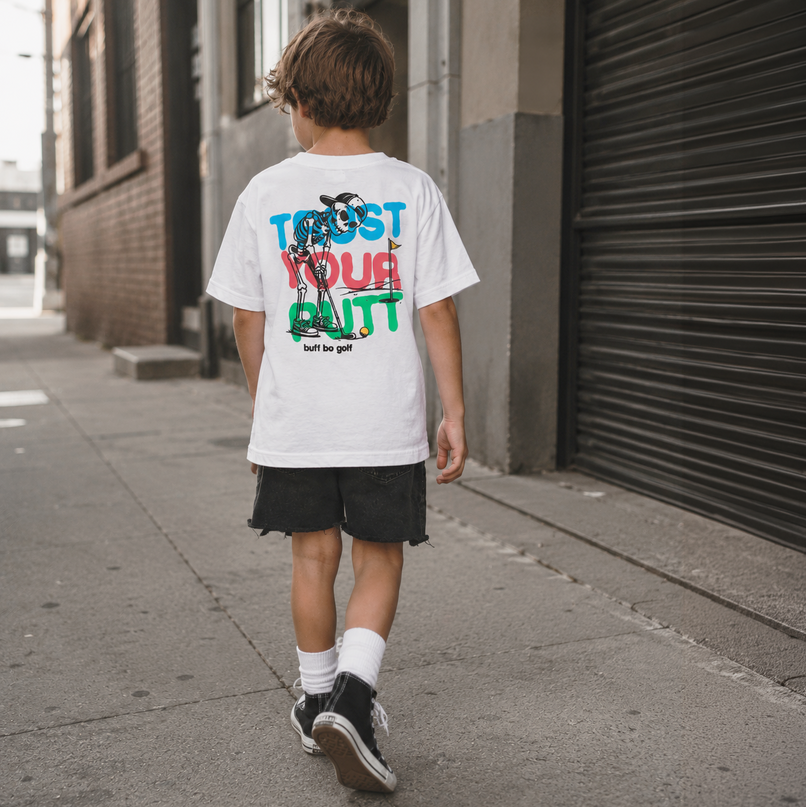 A child viewed from behind walking on a city sidewalk, wearing a white graphic T-shirt with large multicolor text and artwork on the back, black shorts, white socks, and black sneakers.