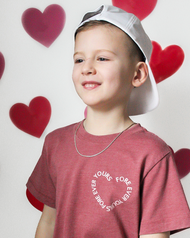 Child wearing a red t-shirt with text and a white cap, standing against a wall with heart shapes.