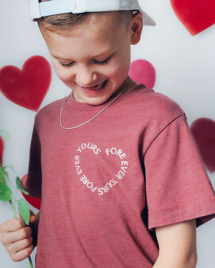 Child wearing a red shirt with text, holding a rose in front of heart-shaped balloons.