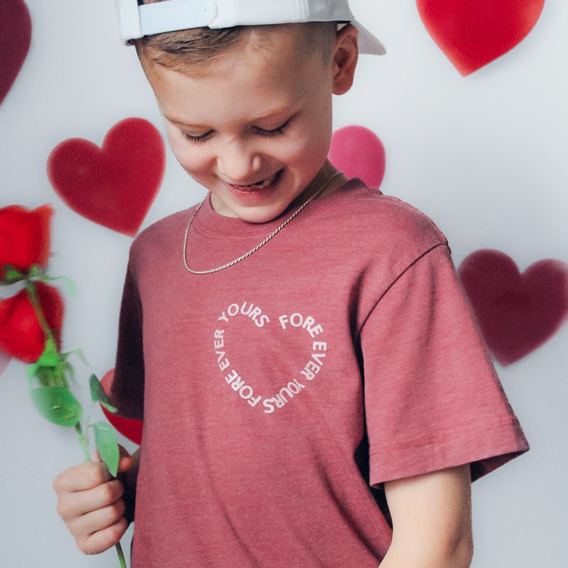 Child wearing a red shirt with text, holding a rose in front of heart-shaped balloons.