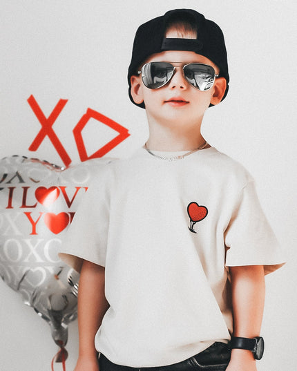 Child wearing a white t-shirt with heart designs and black pants, standing next to a heart-shaped balloon with 'I love you' text.