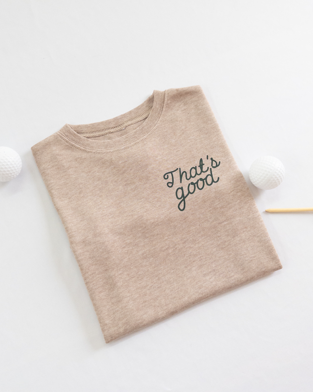 Tan kids golf t-shirt featuring dark green script text reading 'That's good,' styled flat lay with two golf balls and a wooden tee, playful and casual golf apparel for young players.