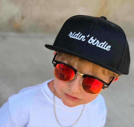 Child wearing a black cap with 'ridin' birdie', white shirt with 'GOLF' text, and red sunglasses.