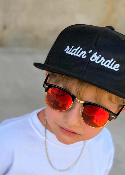 Child wearing a black cap with 'ridin' birdie', white shirt with 'GOLF' text, and red sunglasses.