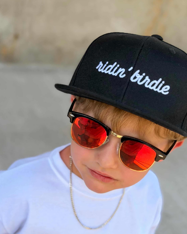 Child wearing a black cap with 'ridin' birdie', white shirt with 'GOLF' text, and red sunglasses.